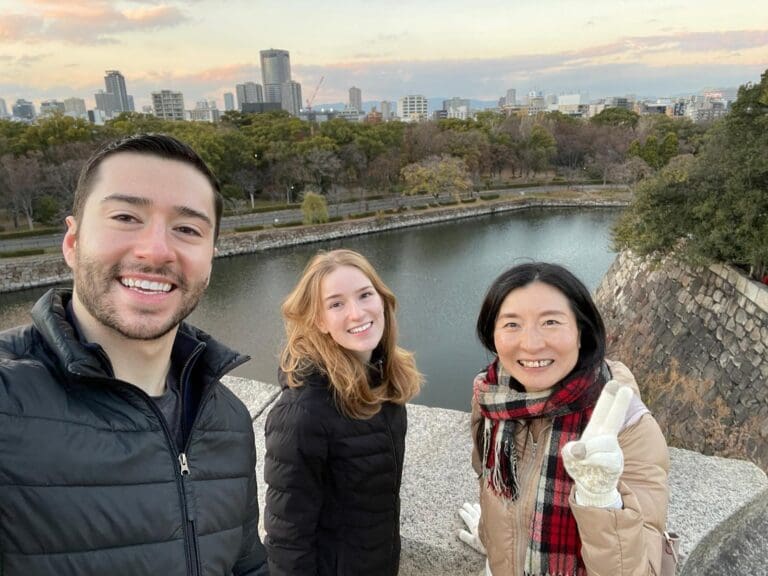 photo-with-amanda-at-the-osaka-castle Three individuals posing for a selfie at a scenic location near a body of water, with a city skyline in the background.