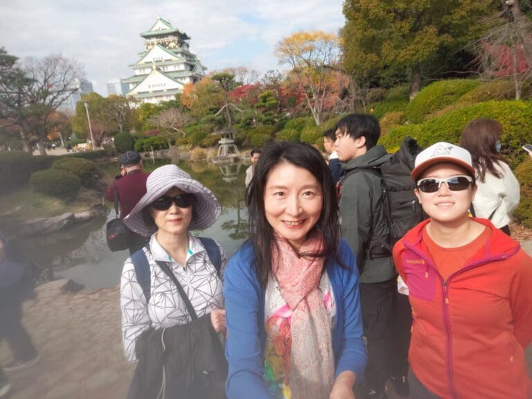 photo-with-jane-and-mun-at-the-osaka-castle Three individuals posing for a selfie at a scenic location near a body of water, with a city skyline in the background.