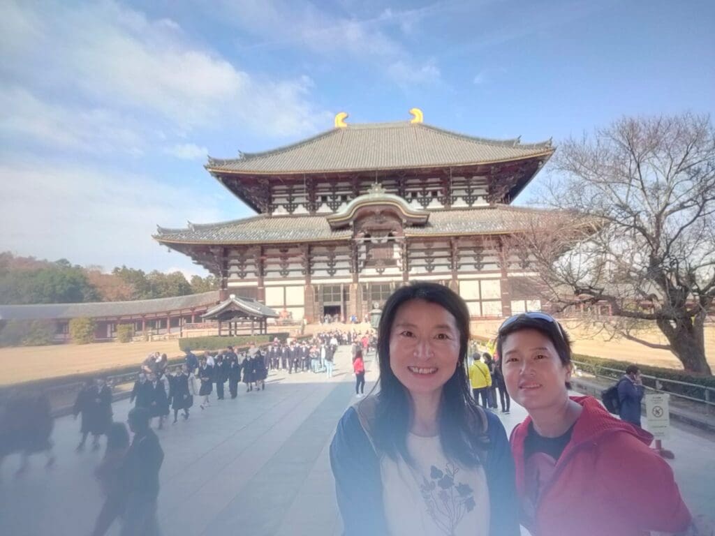 Two individuals posing in front of Todai-ji Temple, a historic Buddhist temple in Nara, Japan, with visitors in the background.