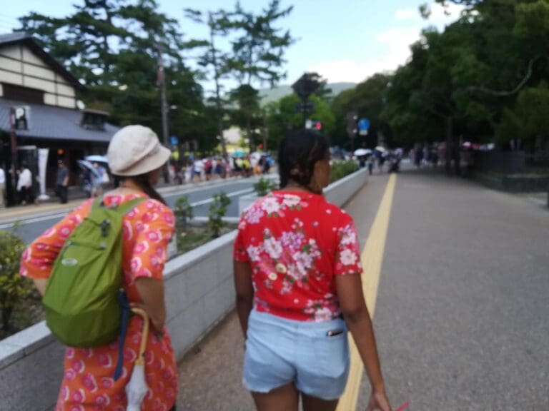 dav-4 Two women walking along a pathway in Nara, wearing colorful floral shirts and shorts, with trees and people in the background.
