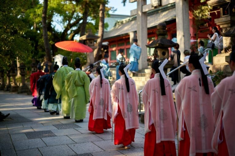 8 Must-See Festivals in Osaka: Culture, Food, and Local Fun 3 A procession of people in traditional Japanese clothing, including pink and red garments, walks through a temple courtyard with stone lanterns and a red umbrella—a vibrant scene reminiscent of festivals in Osaka under bright daylight.