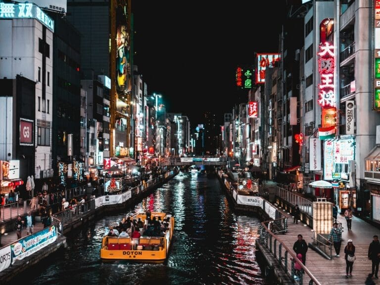 A brightly lit urban canal at night showcases the vibrant nightlife in Osaka, with a yellow sightseeing boat, neon signs on tall buildings, and people strolling along both sides of the water.