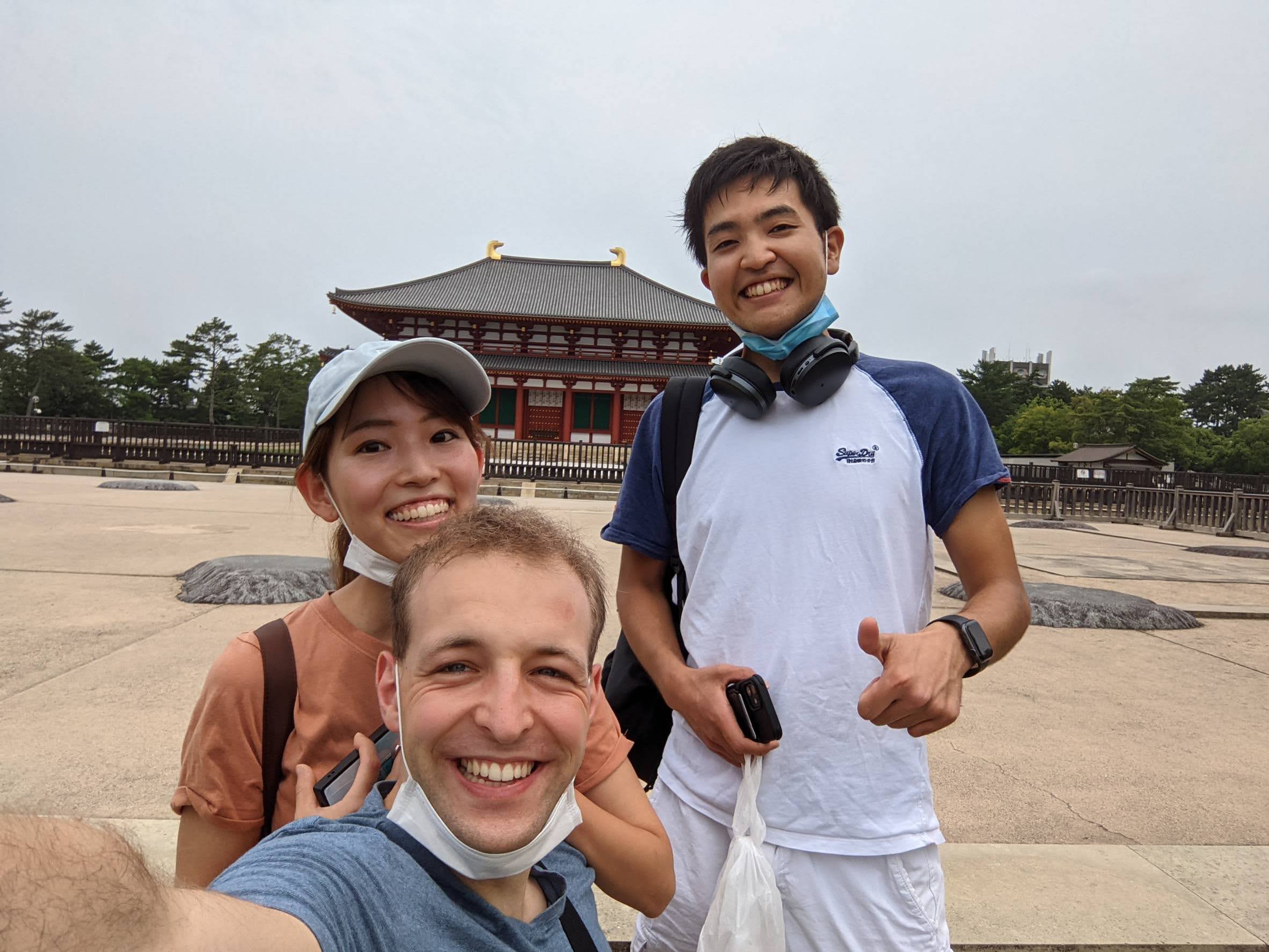 Three people smiling and posing for a selfie in front of a traditional Japanese temple in Osaka, all wearing casual clothes and face masks around their necks. The temple features a curved roof and red accents, capturing the spirit of Kansai local tours.