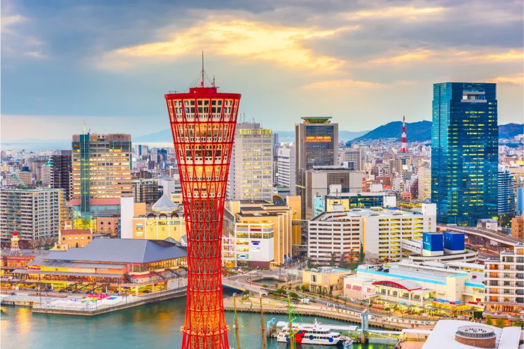 A cityscape of Kobe, Japan at sunset, featuring the red Kobe Port Tower in the foreground, surrounded by modern buildings, waterfront, and distant mountains under a cloudy sky.