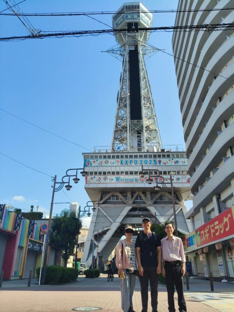photo-with-edward-family Three individuals standing in front of the Tsutenkaku Tower in Osaka, Japan, on a clear day.
