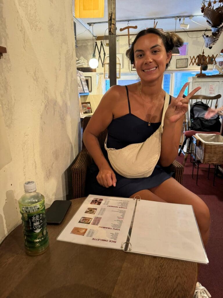 Young woman sitting at a table with a menu, holding a peace sign, and a bottle of green tea nearby.