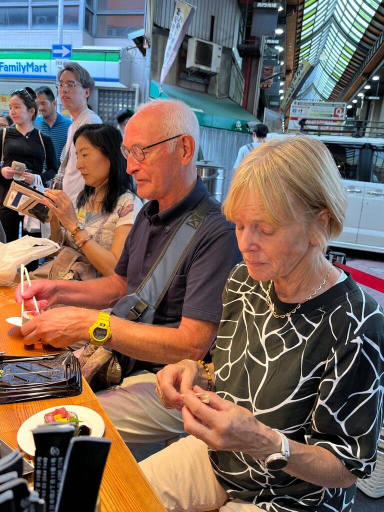 Two individuals seated at a wooden table engaged in an activity, with others in the background at a market.