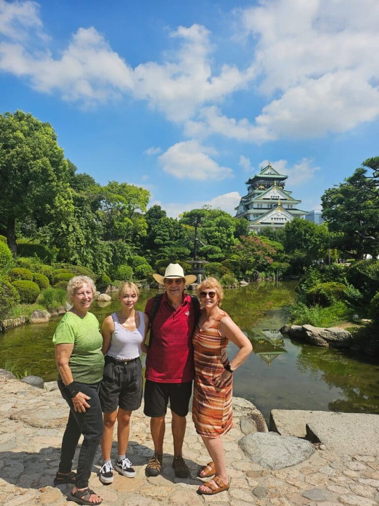 photo-with-shawn-at-the-osaka-castle Group of four people posing in front of Osaka Castle surrounded by greenery and a pond.