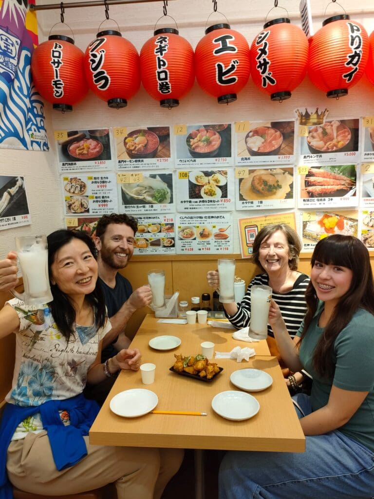 Group of four people raising glasses in a restaurant with Japanese lanterns and menu items in the background.