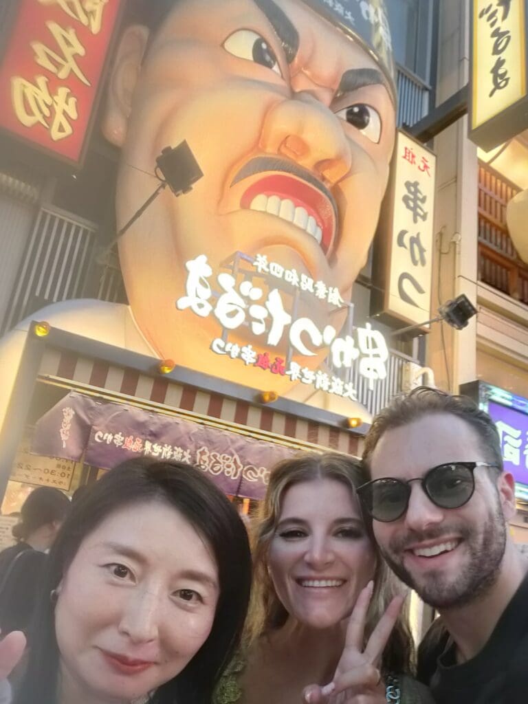 Three individuals posing in front of a large, colorful sign featuring a dramatic face at a restaurant.