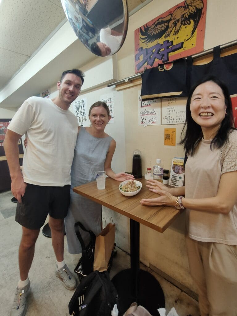 Two guests and a local woman smiling at a table with a bowl of Yakisoba in a casual dining setting.