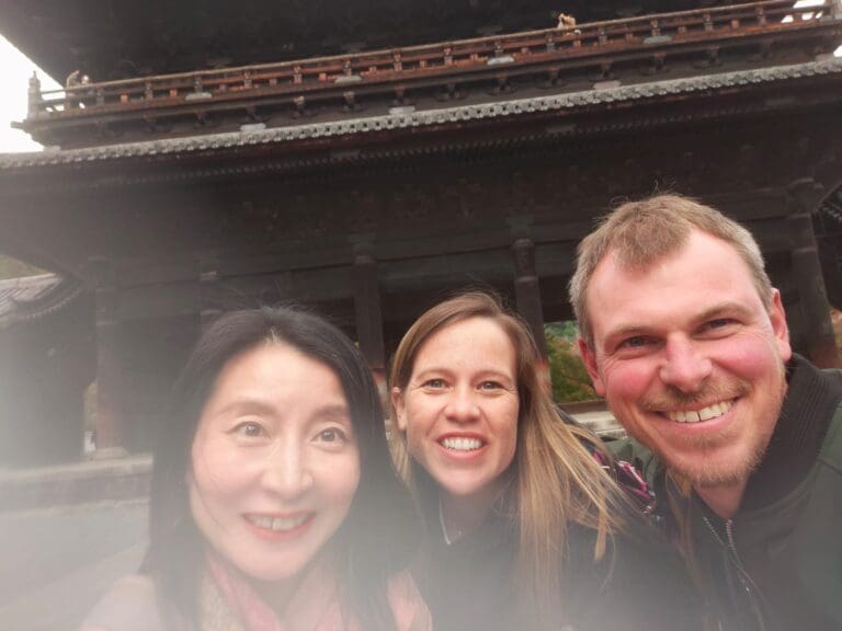 Three individuals posing for a selfie in front of a traditional Japanese temple structure with intricate wooden details.